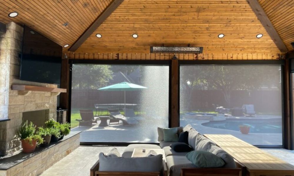 <span class="uk-h4">Covered outdoor living space with a wood-paneled ceiling, stone fireplace and TV on the left, and a large sectional sofa facing motorized shades that look out to a sunny backyard with a pool, lounge chairs, and a green patio umbrella.</span>