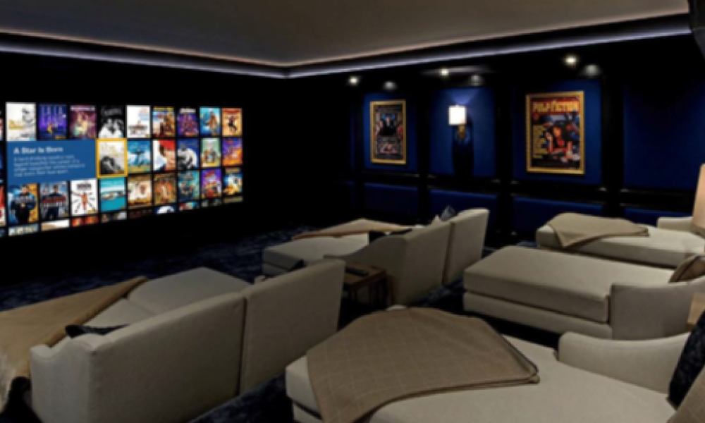 Cozy home theater with several light-colored reclining seats facing a wall-mounted screen of movie covers, dark blue walls, and framed posters.